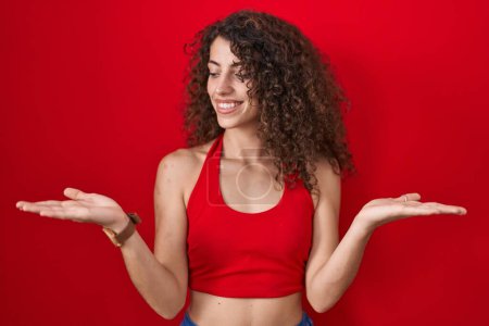 Foto de Hispanic woman with curly hair standing over red background smiling showing both hands open palms, presenting and advertising comparison and balance - Imagen libre de derechos