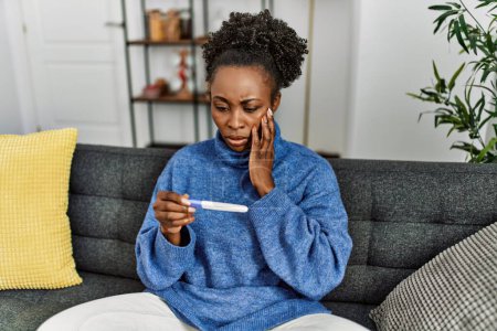 Foto de African american woman holding pregnancy test with worried expression at home - Imagen libre de derechos