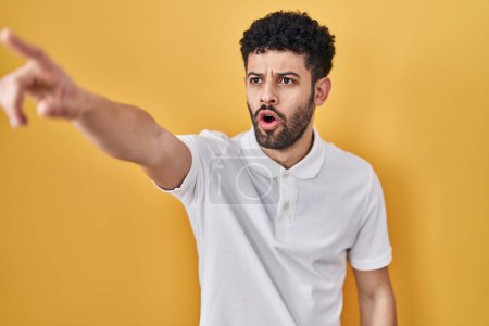 Téléchargez les photos : Arabe debout sur fond jaune pointant avec le doigt surpris devant, bouche ouverte expression étonnante, quelque chose sur le devant - en image libre de droit