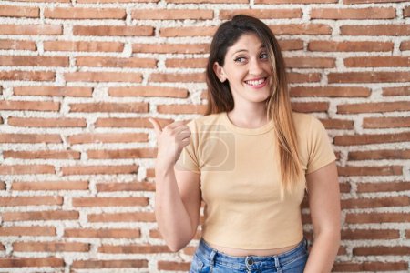 Foto de Young brunette woman standing over bricks wall smiling with happy face looking and pointing to the side with thumb up. - Imagen libre de derechos