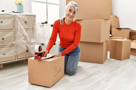 Photo for Middle age grey-haired woman smiling confident packing books package at new home - Royalty Free Image