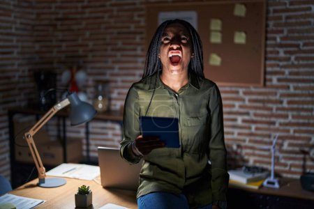 Foto de Joven afroamericano con trenzas trabajando en la oficina por la noche enojado y loco gritando frustrado y furioso, gritando con ira. rabia y concepto agresivo. - Imagen libre de derechos