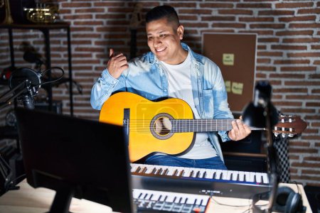 Photo for Hispanic young man playing classic guitar at music studio pointing to the back behind with hand and thumbs up, smiling confident - Royalty Free Image