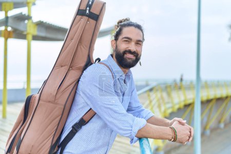 Téléchargez les photos : Jeune musicien hispanique tenant un étui de guitare appuyé sur la balustrade au bord de la mer - en image libre de droit