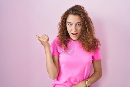 Photo for Young caucasian woman standing over pink background surprised pointing with hand finger to the side, open mouth amazed expression. - Royalty Free Image