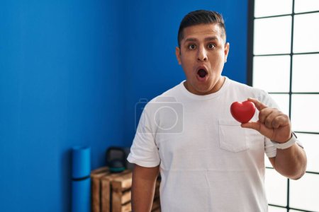 Photo for Hispanic young man holding red heart at gym scared and amazed with open mouth for surprise, disbelief face - Royalty Free Image