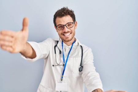 Photo for Young hispanic man wearing doctor uniform and stethoscope looking at the camera smiling with open arms for hug. cheerful expression embracing happiness. - Royalty Free Image