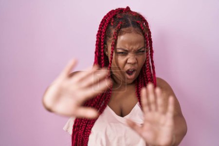 Téléchargez les photos : Femme afro-américaine aux cheveux tressés debout sur fond rose faisant geste d'arrêt avec les mains paumes de main, expression de colère et de frustration - en image libre de droit