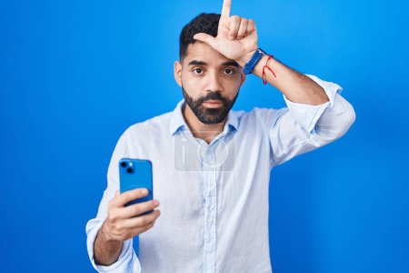 Photo for Hispanic man with beard using smartphone typing message making fun of people with fingers on forehead doing loser gesture mocking and insulting. - Royalty Free Image
