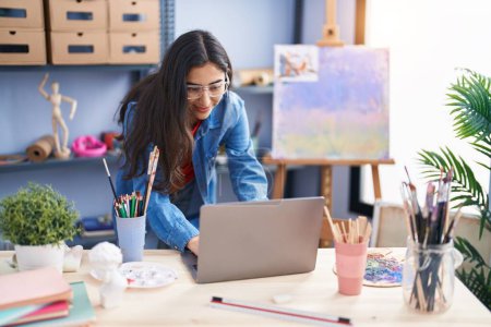 Téléchargez les photos : Young hispanic girl artist smiling confident using laptop at art studio - en image libre de droit