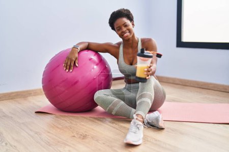 Photo for African american woman drinking smoothie sitting on yoga mat at sport center - Royalty Free Image