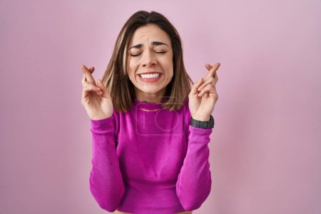 Téléchargez les photos : Hispanic woman standing over pink background gesturing finger crossed smiling with hope and eyes closed. luck and superstitious concept. - en image libre de droit