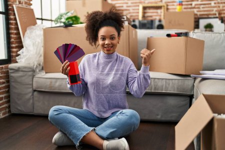 Photo for Young african american woman moving to a new home choosing colors pointing thumb up to the side smiling happy with open mouth - Royalty Free Image