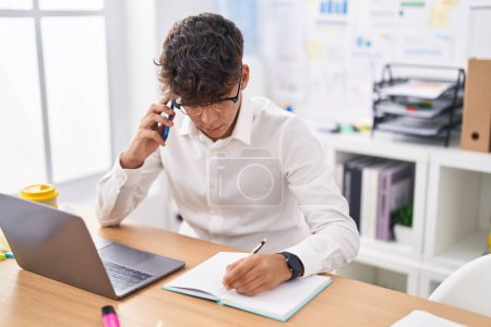 Téléchargez les photos : Young hispanic teenager business worker talking on smartphone writing on notebook at office - en image libre de droit