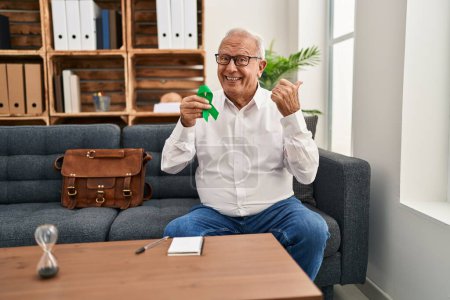 Photo for Senior therapist with grey hair holding support green ribbon for mental health awareness pointing thumb up to the side smiling happy with open mouth - Royalty Free Image
