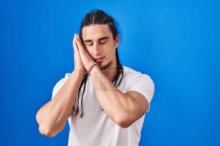 Photo for Hispanic man with long hair standing over blue background sleeping tired dreaming and posing with hands together while smiling with closed eyes. - Royalty Free Image