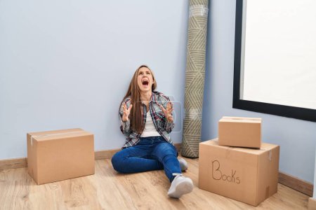 Foto de Young blonde woman sitting on the floor moving to a new home crazy and mad shouting and yelling with aggressive expression and arms raised. frustration concept. - Imagen libre de derechos