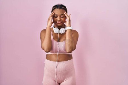 Foto de Mujer afroamericana con trenzas que usan ropa deportiva y auriculares con la mano en la cabeza, dolor de cabeza porque el estrés. padecer migraña. - Imagen libre de derechos