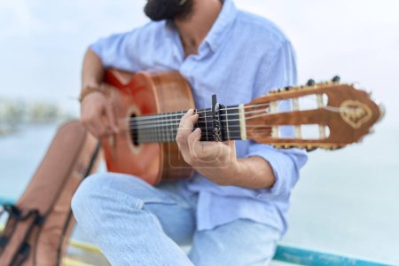 Téléchargez les photos : Jeune homme hispanique musicien jouant de la guitare classique assis sur la balustrade au bord de la mer - en image libre de droit