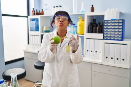 Téléchargez les photos : Femme hispanique travaillant au laboratoire scientifique avec pomme regardant la caméra souffler un baiser étant belle et sexy. expression d'amour. - en image libre de droit