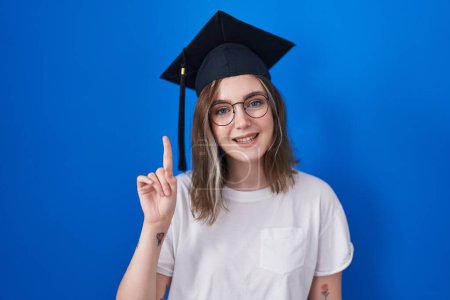 Foto de Mujer rubia caucásica con gorra de graduación mostrando y apuntando hacia arriba con el dedo número uno mientras sonríe confiado y feliz. - Imagen libre de derechos