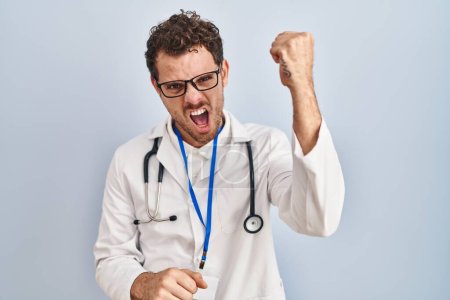 Photo for Young hispanic man wearing doctor uniform and stethoscope angry and mad raising fist frustrated and furious while shouting with anger. rage and aggressive concept. - Royalty Free Image