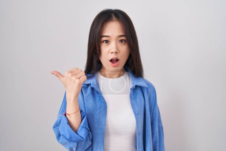 Foto de Joven mujer china de pie sobre fondo blanco sorprendido señalando con el dedo de la mano a un lado, boca abierta expresión asombrada. - Imagen libre de derechos