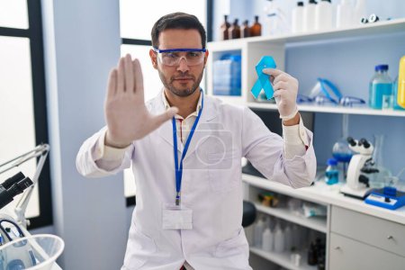 Téléchargez les photos : Jeune homme hispanique avec barbe travaillant au laboratoire scientifique tenant un ruban bleu à la main ouverte faisant signe d'arrêt avec expression sérieuse et confiante, geste de défense - en image libre de droit