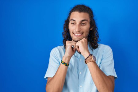 Téléchargez les photos : Jeune homme hispanique debout sur fond bleu riant nerveux et excité avec les mains sur le menton regardant vers le côté - en image libre de droit