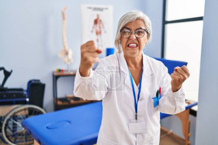 Téléchargez les photos : Middle age woman with grey hair working at pain recovery clinic angry and mad raising fists frustrated and furious while shouting with anger. rage and aggressive concept. - en image libre de droit