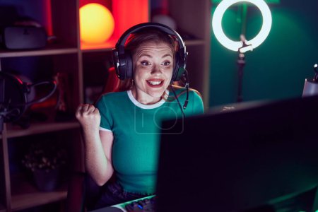 Téléchargez les photos : Femme rousse jouant à des jeux vidéo criant fier, célébrant la victoire et le succès très excité avec les bras levés - en image libre de droit
