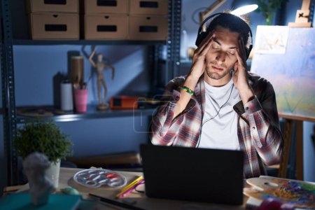 Téléchargez les photos : Jeune homme hispanique assis au studio d'art avec ordinateur portable tard dans la nuit avec la main sur la tête pour la douleur à la tête parce que le stress. migraine souffrant. - en image libre de droit