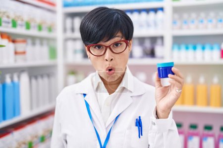 Photo for Young asian woman with short hair working at pharmacy drugstore holding cream jar scared and amazed with open mouth for surprise, disbelief face - Royalty Free Image