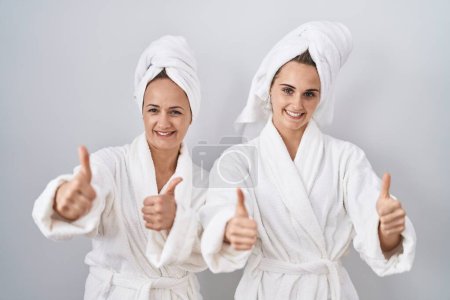 Téléchargez les photos : Femme d'âge moyen et fille portant un peignoir blanc et une serviette approuvant faire un geste positif avec la main, le pouce levé souriant et heureux pour le succès. geste gagnant. - en image libre de droit