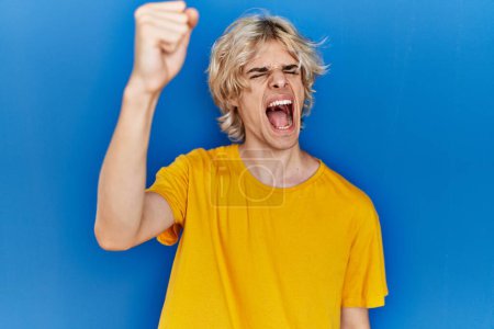 Photo for Young modern man standing over blue background angry and mad raising fist frustrated and furious while shouting with anger. rage and aggressive concept. - Royalty Free Image