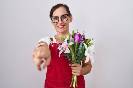 Téléchargez les photos : Middle age brunette woman wearing apron working at florist shop holding bouquet pointing to you and the camera with fingers, smiling positive and cheerful - en image libre de droit