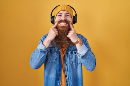 Téléchargez les photos : Caucasian man with long beard listening to music using headphones smiling with open mouth, fingers pointing and forcing cheerful smile - en image libre de droit
