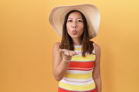 Foto de Mujer china de mediana edad con sombrero de verano sobre fondo amarillo mirando a la cámara soplando un beso con la mano en el aire siendo encantador y sexy. expresión de amor. - Imagen libre de derechos