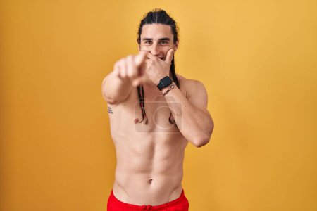 Téléchargez les photos : Hispanic man with long hair standing shirtless over yellow background laughing at you, pointing finger to the camera with hand over mouth, shame expression - en image libre de droit