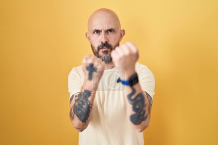 Téléchargez les photos : Homme hispanique avec des tatouages debout sur fond jaune prêt à se battre avec geste de défense poing, visage en colère et bouleversé, peur du problème - en image libre de droit