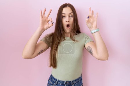Photo for Beautiful brunette woman standing over pink background looking surprised and shocked doing ok approval symbol with fingers. crazy expression - Royalty Free Image