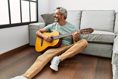 Photo for Middle age grey-haired man playing classical guitar sitting on floor at home - Royalty Free Image