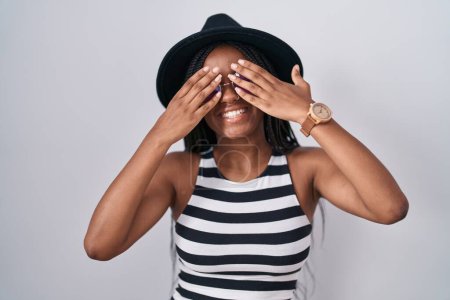 Téléchargez les photos : Jeune afro-américain avec des tresses portant un chapeau et des lunettes de soleil couvrant les yeux avec les mains souriantes gaies et drôles. concept aveugle. - en image libre de droit