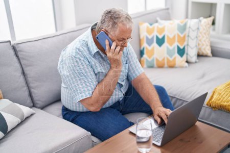 Foto de Hombre de pelo gris de mediana edad hablando en el teléfono inteligente usando el ordenador portátil sentado en el sofá en casa - Imagen libre de derechos