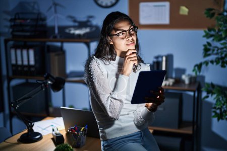 Photo for Young brazilian woman using touchpad at night working at the office looking confident at the camera smiling with crossed arms and hand raised on chin. thinking positive. - Royalty Free Image