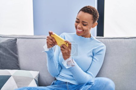 Photo for African american woman playing video game sitting on sofa at home - Royalty Free Image