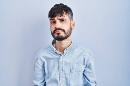 Foto de Joven hombre hispano con barba de pie sobre fondo azul despistado y confundido con los brazos abiertos, sin concepto de idea. - Imagen libre de derechos