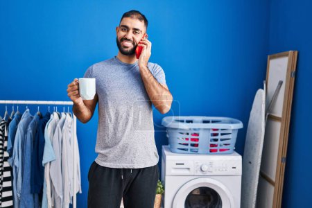 Photo for Young hispanic man talking on smartphone drinking coffee waiting for washing machine at laundry room - Royalty Free Image