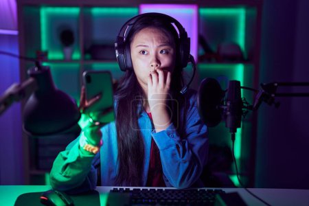 Foto de Joven mujer asiática jugando videojuegos con smartphone buscando estresado y nervioso con las manos en la boca mordiendo las uñas. problema de ansiedad. - Imagen libre de derechos