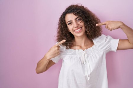 Téléchargez les photos : Femme hispanique avec les cheveux bouclés debout sur fond rose souriant joyeuse montrant et pointant avec les doigts les dents et la bouche. concept de santé dentaire. - en image libre de droit